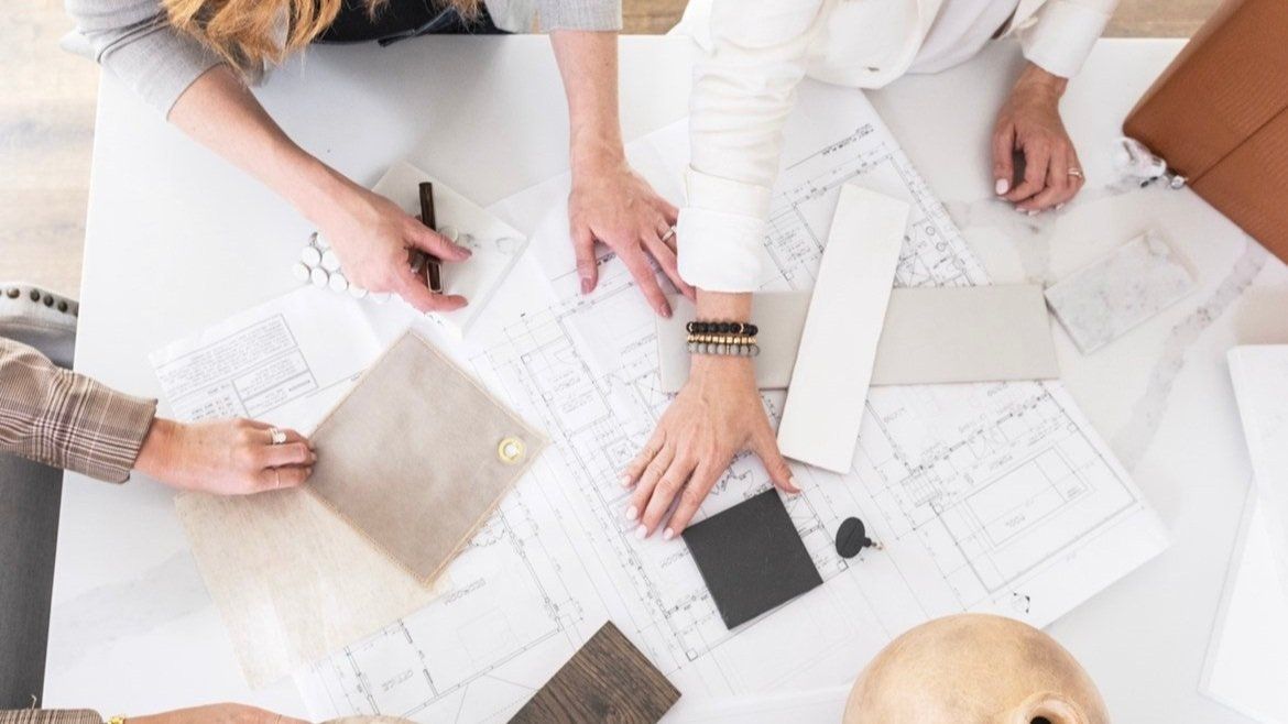 Table with architectural designs, fabrics, tile, and other home build items laid out, with hands reaching in working on the project. 